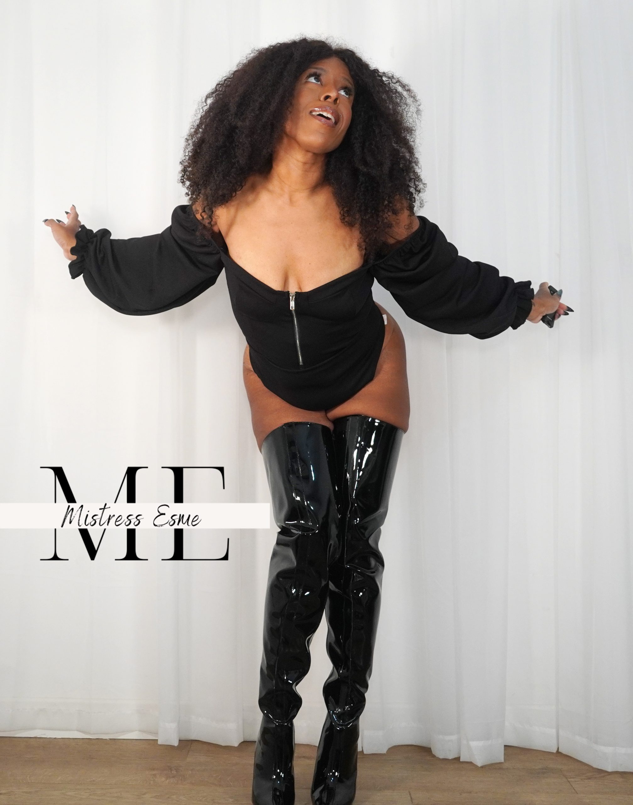 Mistress Esme is in a black bodysuit with long sleeves and black pvc thigh high boots. She has her arms wide open and is looking up with a smile on her face as if she knows something.